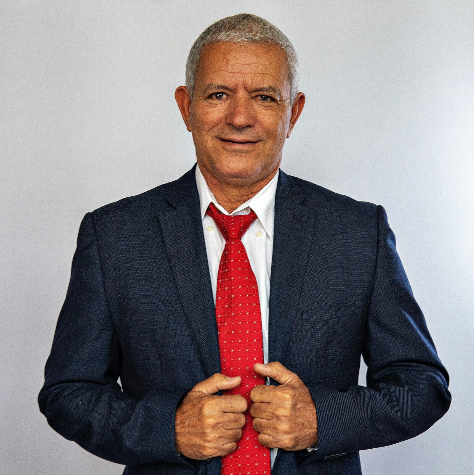 Senior businessman smiling confidently in a suit and red tie against a gray background.